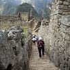 People walk up stone steps surrounded by ancient ruins at Machu Picchu, with green mountains and cloudy skies in the background—an unforgettable highlight of any Brazil-Peru trip.