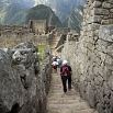 People walk down a narrow stone staircase between ancient stone walls at Machu Picchu, soaking in green mountains and historic ruins—a fabulous trip through breathtaking ancient wonder.