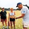 A man in a white outfit and cap holds an object while explaining something to two people outdoors—one in a dark shirt and shorts, the other in a light tank top and shorts. The scene is set in a grassy, open area where details are handled flawlessly.