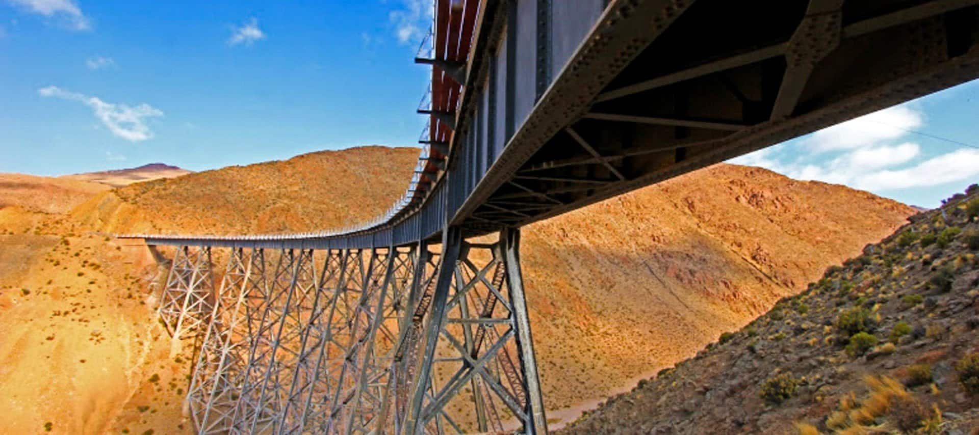 A low-angle view of a metal railway bridge, part of Argentina Train Tours, spans arid, rocky hills under a bright blue sky with some clouds. The structure’s supports and trusses are visible in the foreground.