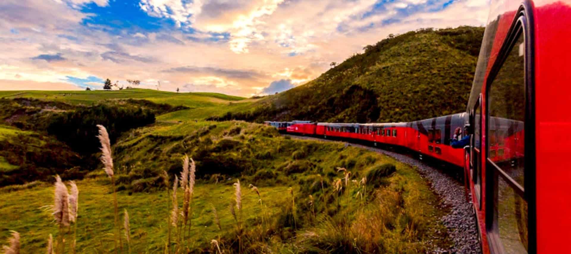 A red train travels along a curving track through green hills under a vibrant, partly cloudy sky at sunset or sunrise—an iconic scene from Ecuador Railway Tours.