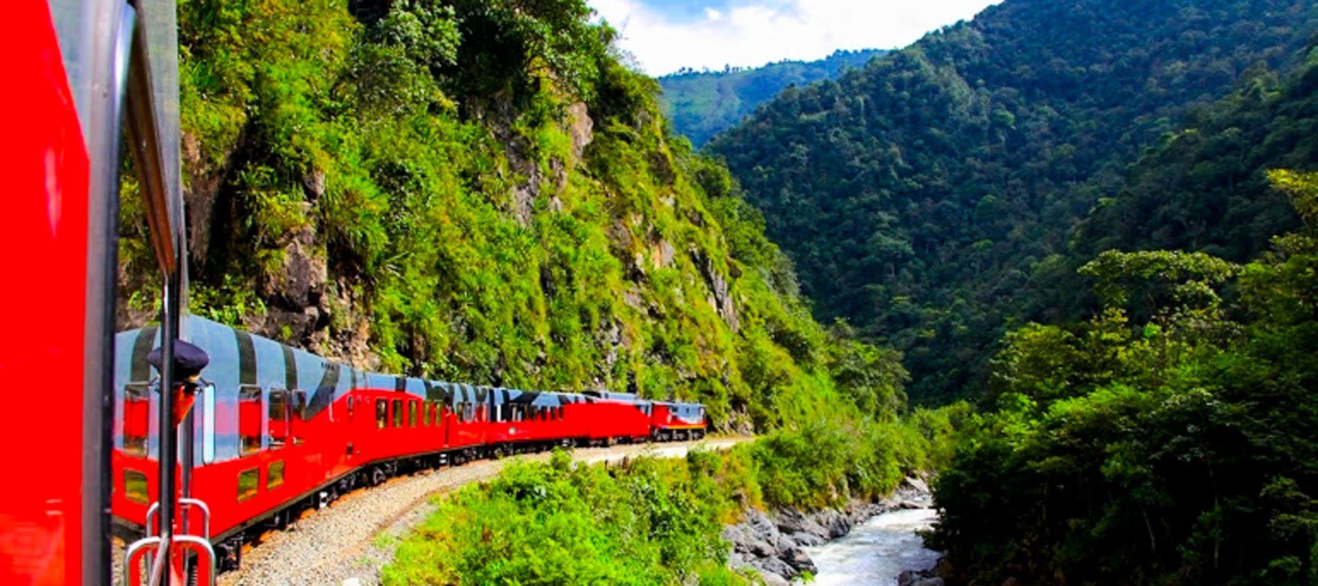A bright red train travels along a winding track beside a river in South America, surrounded by lush green mountains and dense forest under a clear sky.