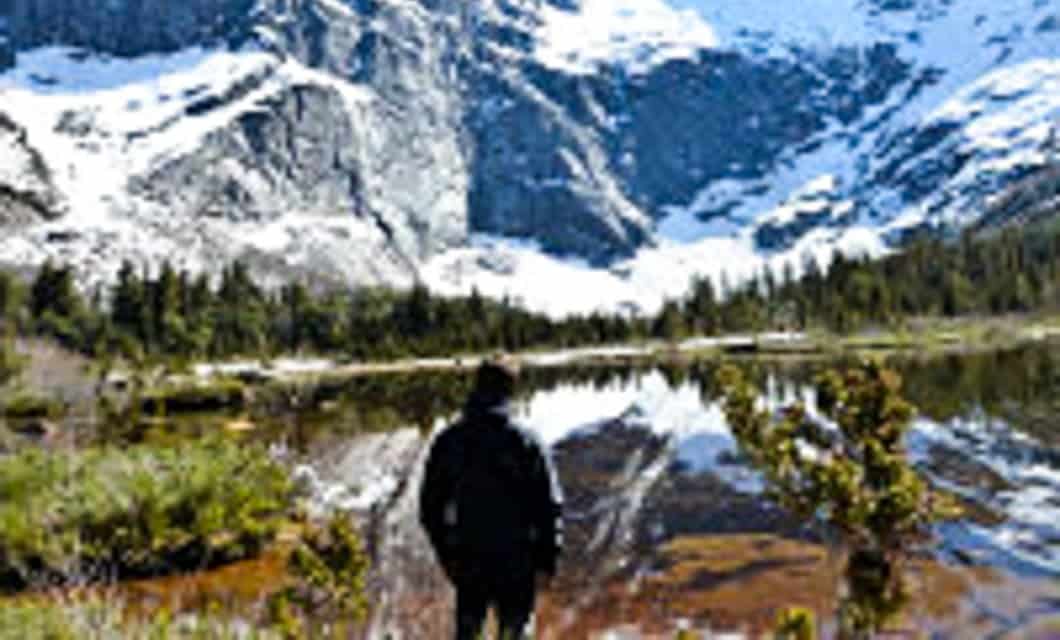 A person stands at the edge of a clear mountain lake near Mitico Puelo Lodge, surrounded by green trees and snow-covered peaks under a blue sky.
