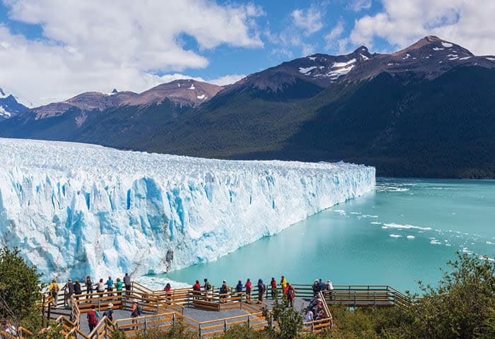 People stand on wooden viewing platforms overlooking a massive blue glacier meeting turquoise water, with forested mountains and a partly cloudy sky beyond—one of the top Argentina attractions and must-see things to do in Argentina.