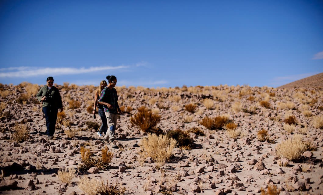 Three people walk through a rocky, arid landscape with scattered dry bushes under a clear blue sky, evoking the rugged beauty near Casa Atacama in a striking desert or semi-desert environment.