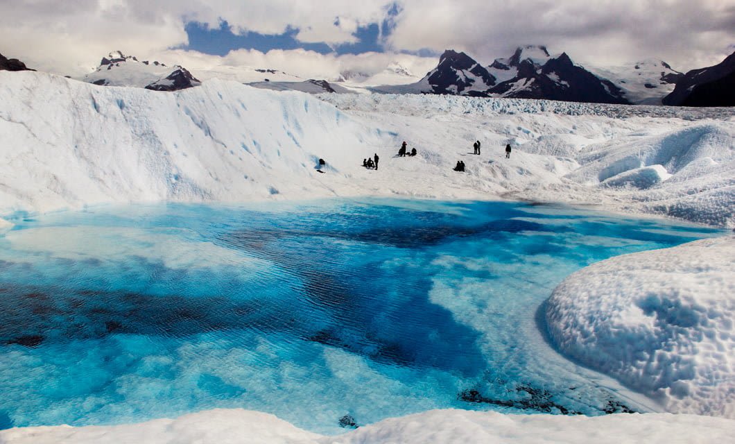 A vivid blue meltwater pool sits surrounded by snow and ice near a Patagonia Lodge, with distant groups of people walking across the glacier; snow-covered mountains rise in the background under a partly cloudy sky.