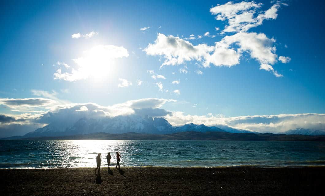 Three people walk along a pebbled lakeshore near Tierra Patagonia, with the sun shining brightly above, mountains shrouded in clouds behind them, and a blue sky dotted with scattered clouds overhead.