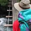 A person wearing a wide-brimmed hat and colorful jacket stands at the edge of a narrow wooden suspension bridge over a rushing river, taking in the travel adventure that awaits on this fabulous trip.