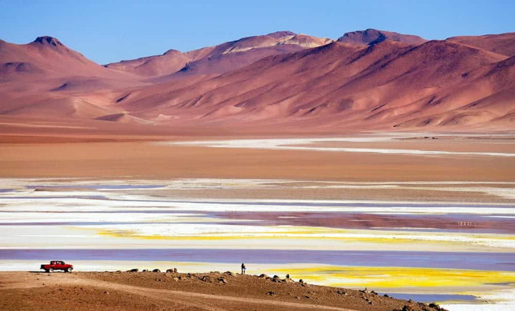A red car parked on a dirt road near Explora Atacama overlooks a colorful, multi-hued salt flat with yellow, white, and purple patches, set against reddish mountains and a clear blue sky.