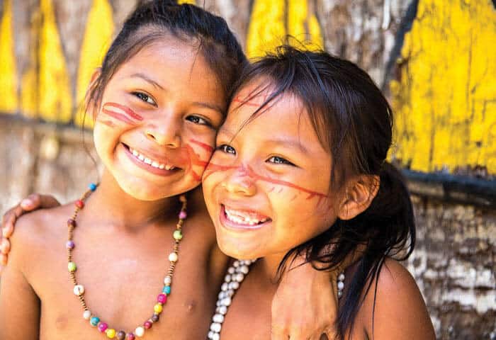Two smiling children with traditional red face paint and beaded necklaces stand close together, arms around each other, in front of a tree with yellow designs—capturing the vibrant spirit of Amazon activities in the Amazon Rainforest.