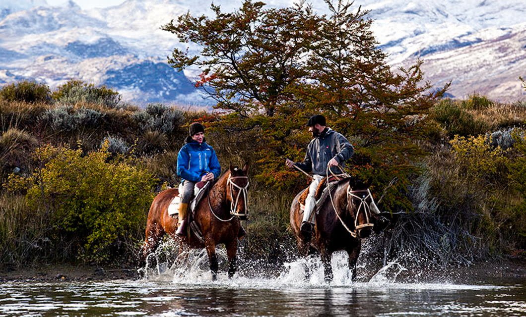 Two people ride horses through a shallow stream at Estancia Cristina, surrounded by autumn foliage and snow-capped mountains in Patagonia, Argentina. Both riders wear jackets and hats, enjoying the scenic outdoor landscape.