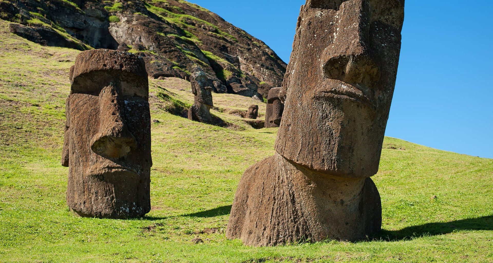 Large stone moai statues stand on a grassy hillside under a clear blue sky on Easter Island, Chile, a UNESCO World Heritage site, with more statues and a rocky hill visible in the background.