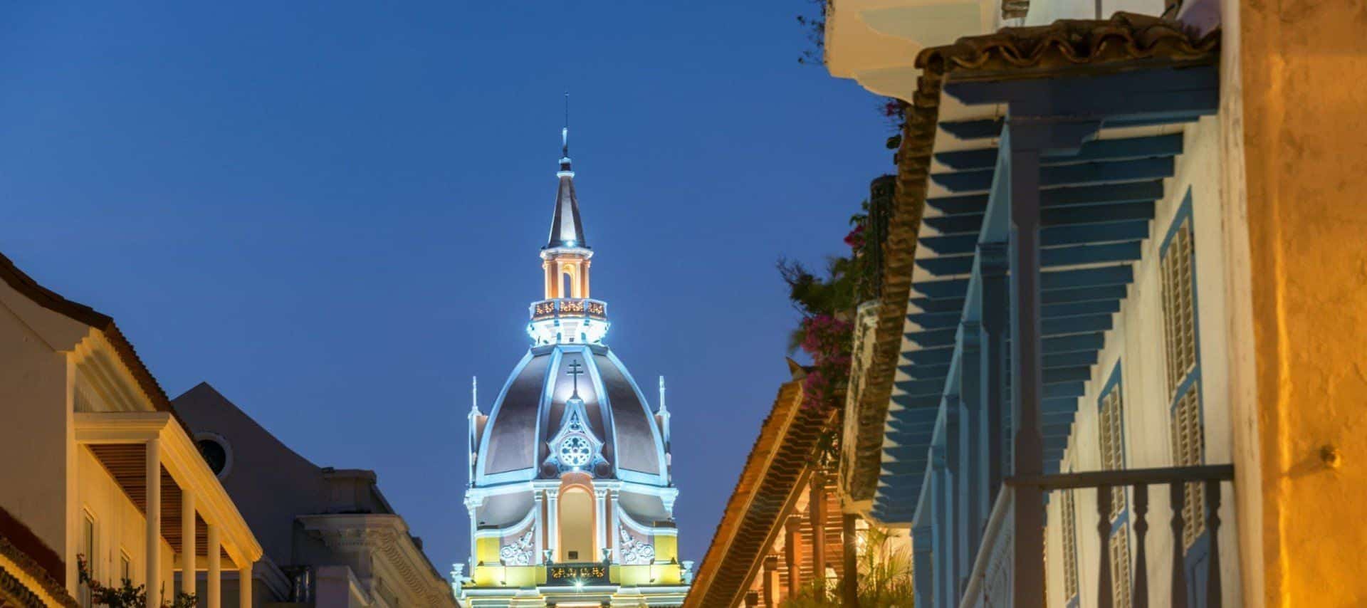 A brightly lit dome of a historic church rises above colorful colonial-style buildings at dusk in Cartagena, Colombia—a UNESCO World Heritage Site in South America. The clear, deep blue sky highlights the stunning architecture.