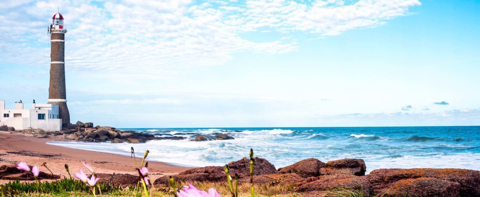 A tall lighthouse stands on a rocky shore near a white building, overlooking waves crashing onto a sandy beach under a partly cloudy blue sky—an inviting scene for Uruguay travel. Pink flowers bloom in the foreground.