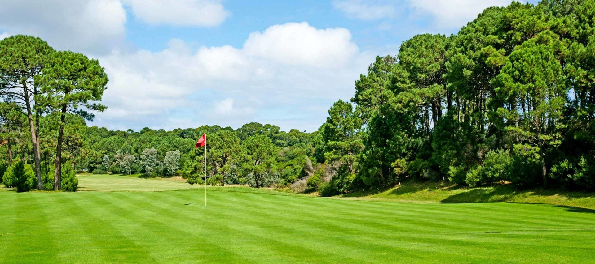 A well-manicured golf course with a red flag on the green, surrounded by lush trees and blue sky with scattered clouds—perfect for unforgettable Uruguay Golf Tours.