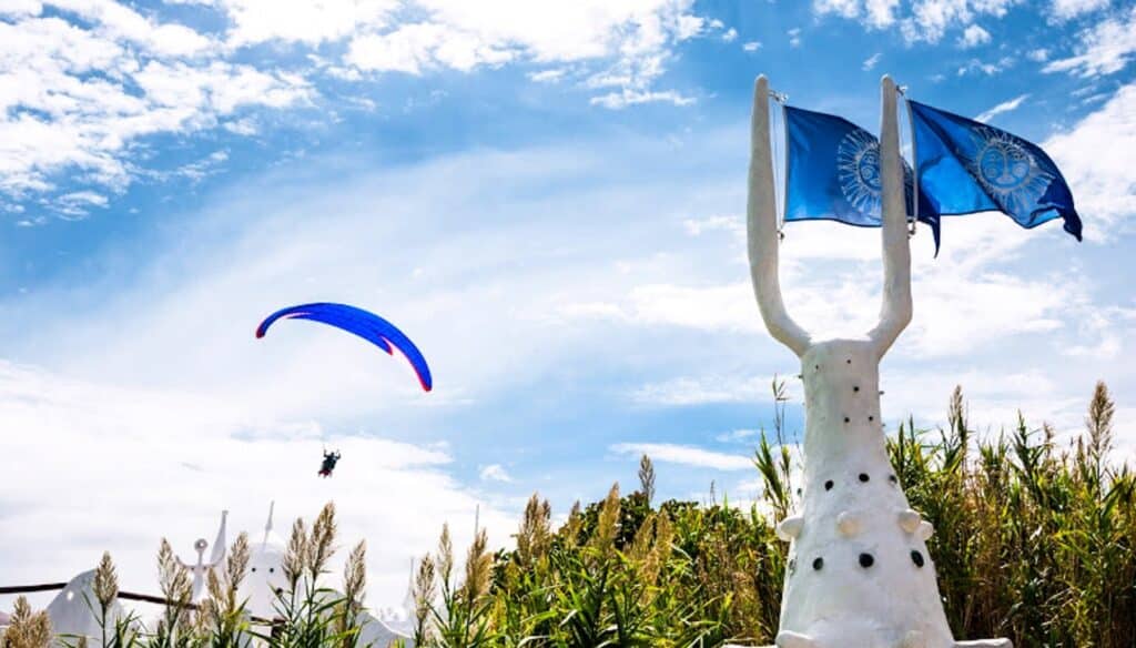 A paraglider with a blue canopy soars through a partly cloudy sky, near a tall white sculpture with two blue flags and lush green plants—an inspiring view to add adventure to your Uruguay packing list.