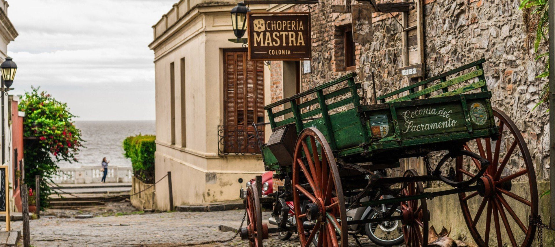 A rustic cobblestone street in Uruguay with an old wooden cart and motorcycle near stone buildings, a Chopería Mastra sign, and water views. Greenery decorates the left side, inviting tours of this UNESCO heritage site. A person stands in the distance.