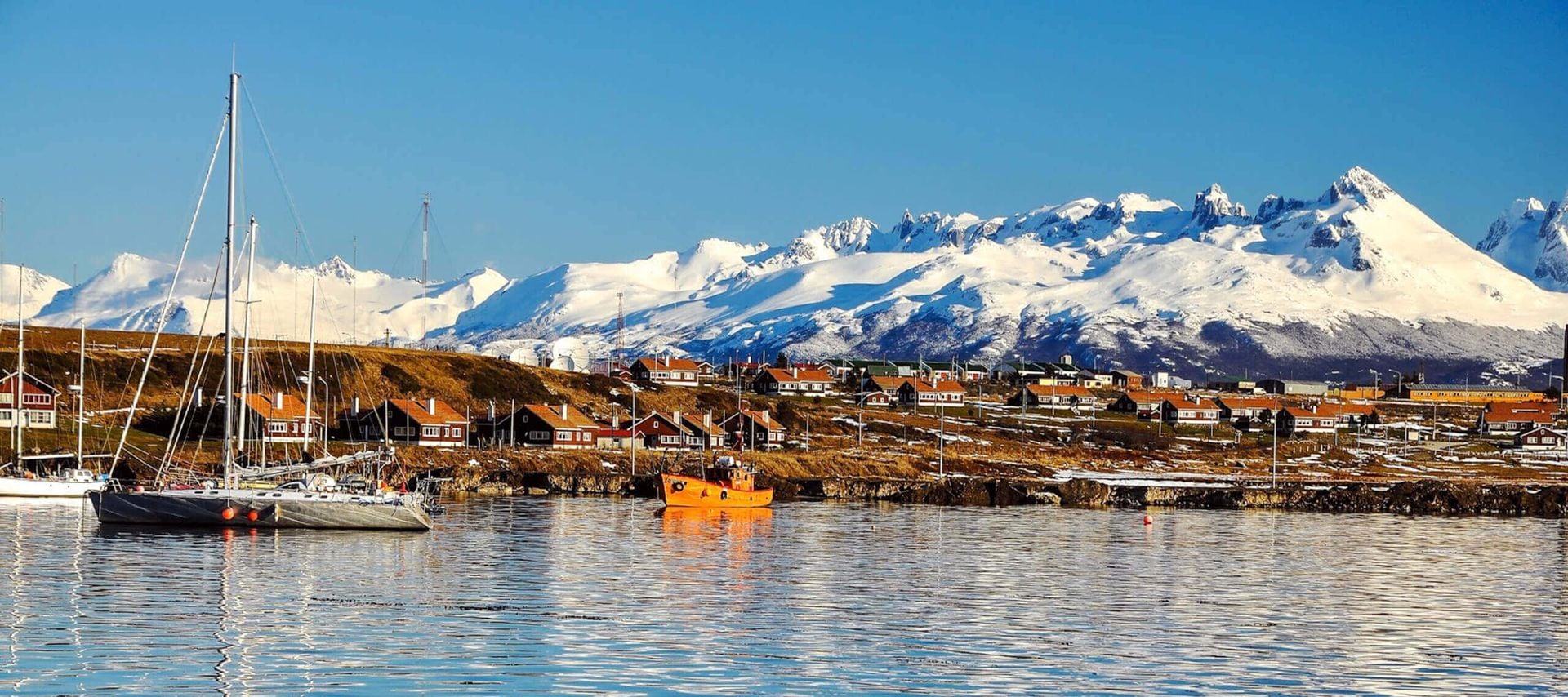 A calm harbor with sailboats and an orange boat, reflecting in the water, sits before a small town with red-roofed buildings and snow-capped mountains—an inviting scene for Ushuaia cruises under a clear blue sky.