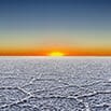 A bright orange sun sets on the horizon above a vast, flat salt desert with cracked white ground, capturing the perfect end to a memorable trip under a clear blue sky gradually fading to yellow near the horizon.