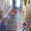 A group of children and adults walk and play on colorful, graffiti-covered outdoor stairs between tall walls filled with street art, creating a wonderful experience as kids climb, sit, or pose on the vibrant steps.