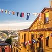 Colorful strings of international flags hang above a narrow street lined with yellow buildings, creating the perfect backdrop for a trip of a lifetime, with a scenic river and cityscape visible under a clear blue sky.