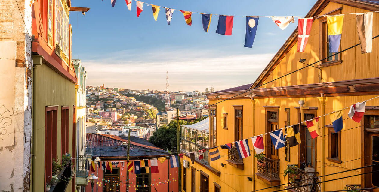 Colorful hillside buildings with vibrant yellow and green facades line a narrow street in Valparaíso, Chile. Strings of international flags hang above, and the cityscape spreads out under a clear sky in the background.