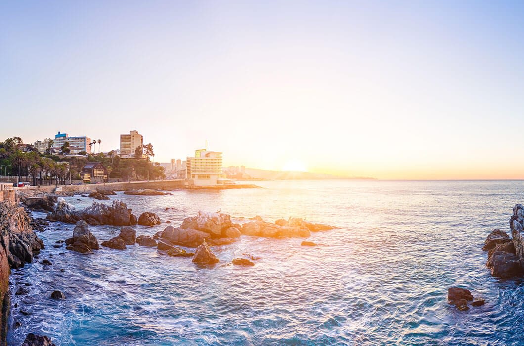 Waves crash against rocky shores under a clear sky at sunrise, with coastal buildings and palm trees in the distance—capturing the beauty of Chile tourism and one of the must-see Chile attractions along its picturesque coastline.