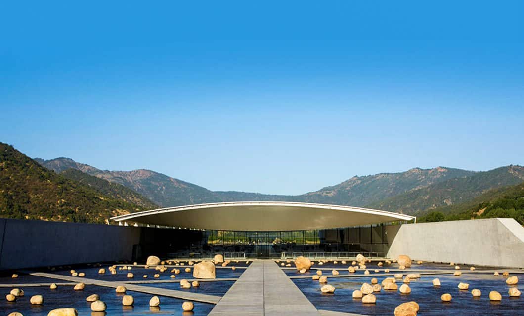 A modern building with a large curved roof sits amid mountains at Viña Vik, a luxury hotel featuring a courtyard with water and scattered rocks under a clear blue sky.