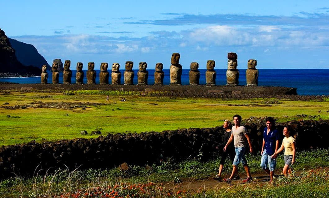 Four people walk along a path in front of the famous row of large Moai statues on grassy Rapa Nui (Easter Island), with the ocean and blue sky in the background.