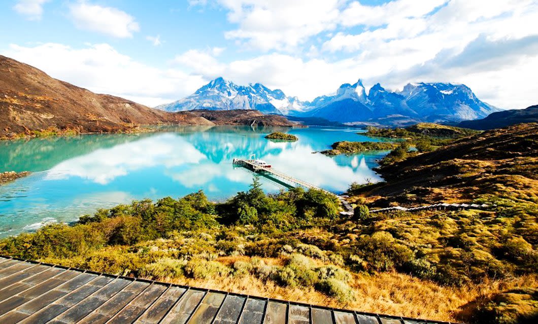 A turquoise lake in Patagonia is surrounded by mountains and lush greenery, with a narrow bridge leading to a small island and snow-capped peaks in the background—an inviting scene for adventure travel.