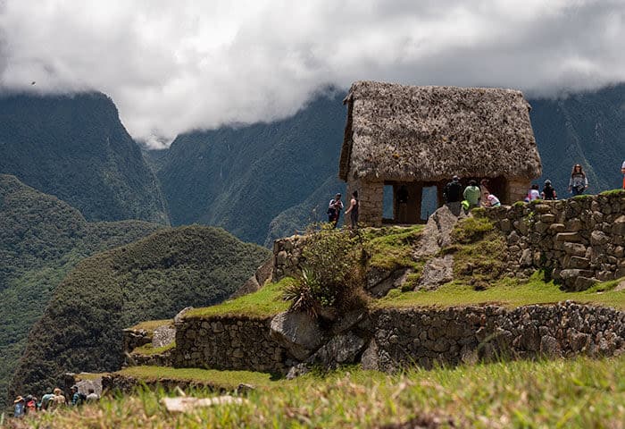 Thatched stone building on a grassy terrace at Machu Picchu, surrounded by visitors and lush green mountains under a cloudy sky—a must-see for anyone exploring things to do while you travel in Peru.