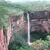 A tall, narrow waterfall cascades down a steep red rock cliff surrounded by dense green vegetation and mist, creating a breathtaking travel experience as it blends into the lush forest below.