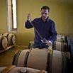 A person in a blue shirt, perhaps on vacation, uses a wine thief to extract liquid from a wooden barrel in a room filled with similar barrels and yellow walls.