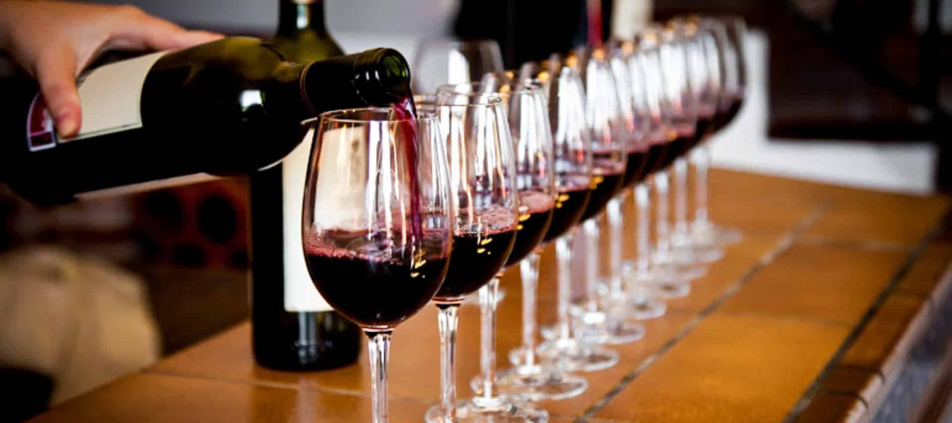 A hand pours red wine from a bottle into one of several wine glasses arranged in a row on a tiled counter, capturing the elegance of Uruguay Wine Tours. A second wine bottle stands in the background.