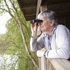 An older man stands on a wooden balcony or deck, enjoying the wonderful service as he looks through binoculars toward a wooded area and a body of water, surrounded by green foliage.