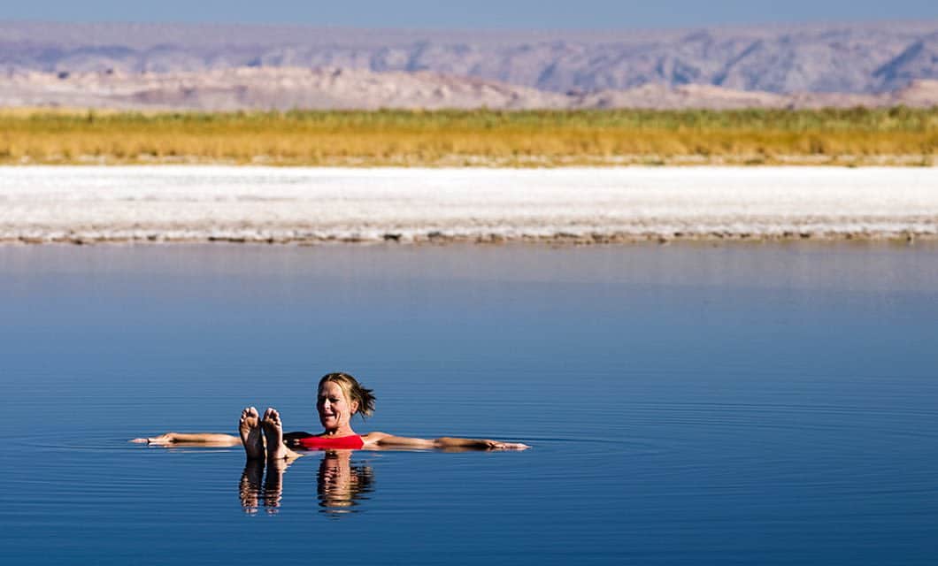 A woman floats on her back in calm, blue water at Tierra Atacama, holding a book. In the background, a strip of grassy land and distant mountains stretch beneath a clear sky near the hotel spa.