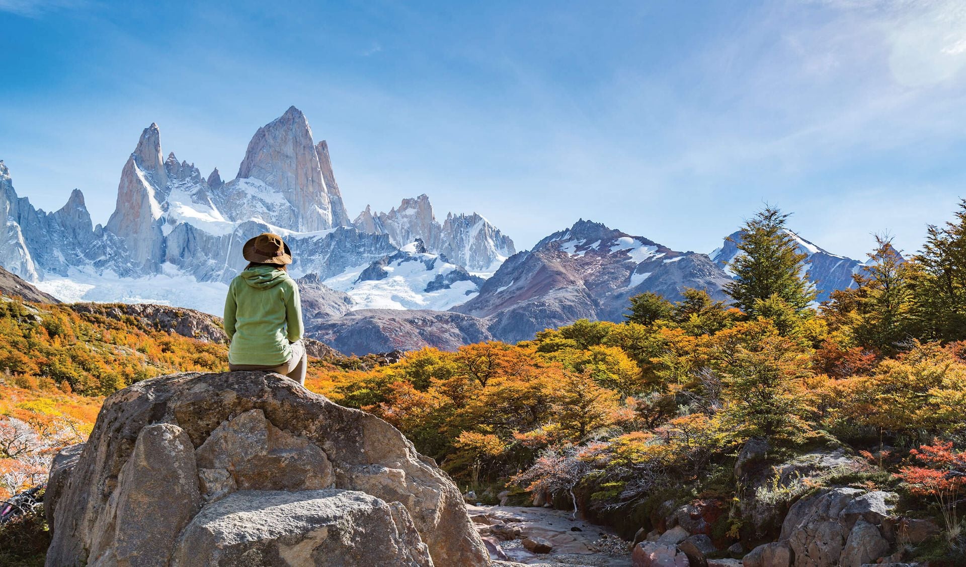 A person in a green jacket and hat sits on a rock, overlooking colorful autumn trees and snow-capped mountain peaks under a bright blue sky—the scene captures the Best Time to Visit Patagonia.