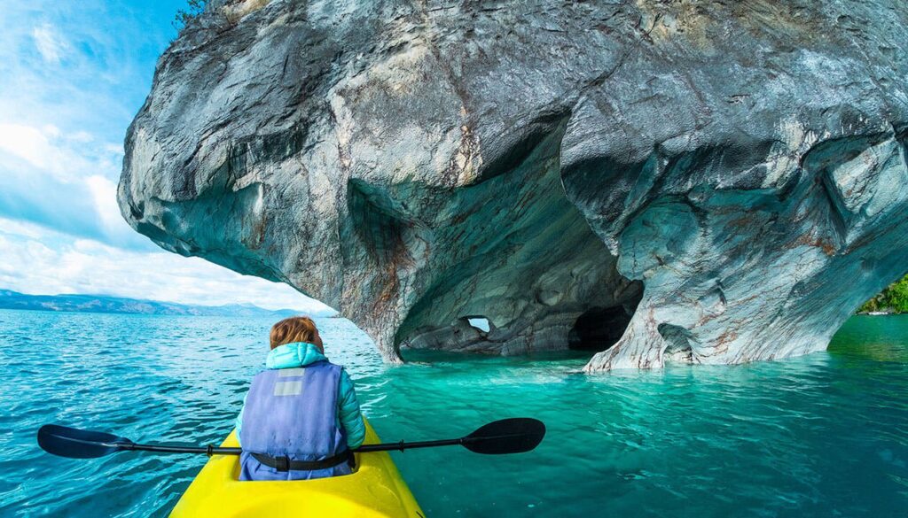 A person in a yellow kayak paddles toward the striking Marble Caves of Patagonia, Chile, where natural arches and patterns rise from turquoise blue water under a partly cloudy sky.