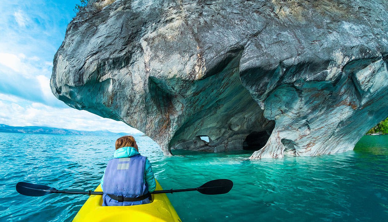 A person in a yellow kayak paddles toward the striking Marble Caves of Patagonia, Chile, where natural arches and patterns rise from turquoise blue water under a partly cloudy sky.