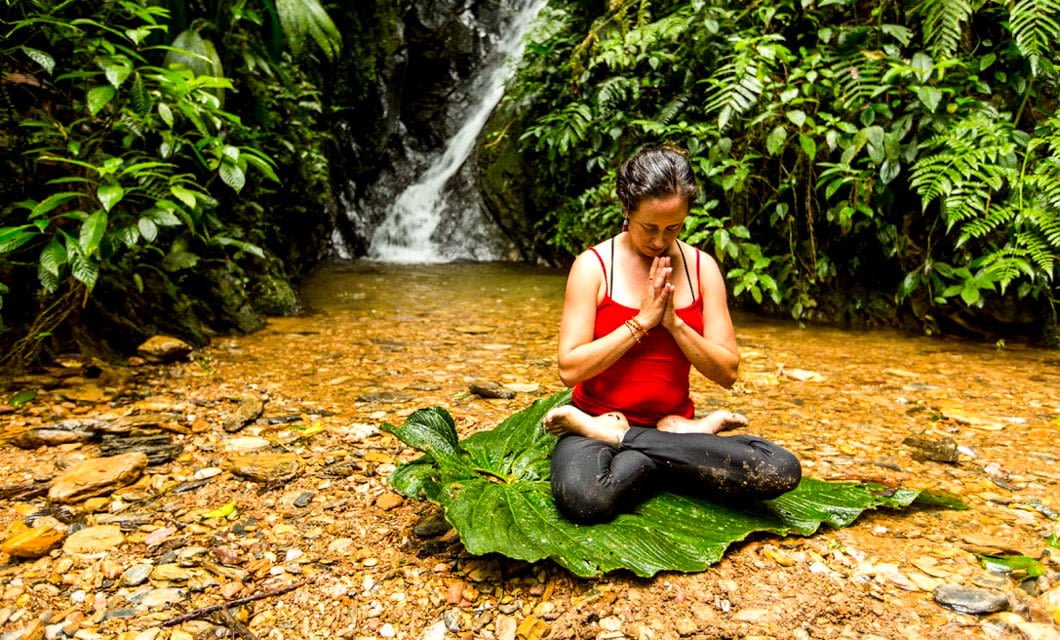 A woman sits cross-legged on a large leaf in a shallow stream, meditating with hands in prayer position. She is surrounded by lush green plants and a waterfall, embodying the tranquil spirit of Mashpi Lodge in Ecuador.