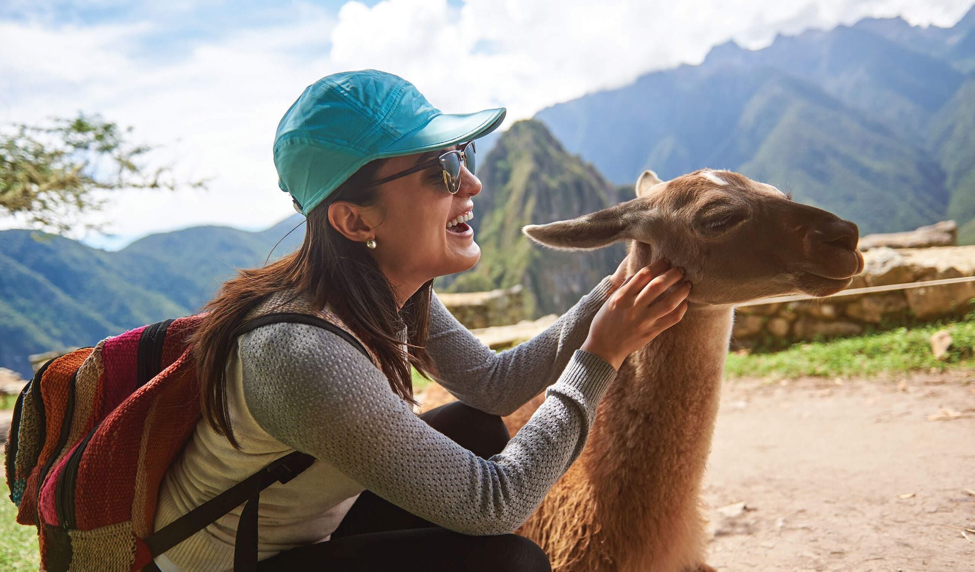 A smiling woman wearing a blue cap, sunglasses, and a colorful backpack pets a llama outdoors at Machu Picchu, with mountains and ancient ruins in the background—one of the top things to do for any travel enthusiast.