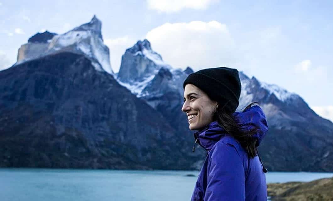 A woman in a purple jacket and black beanie smiles while standing near a lake in Patagonia, with tall, snow-capped mountains in the background under a partly cloudy sky—an ideal scene for adventure travel with Explora Patagonia.