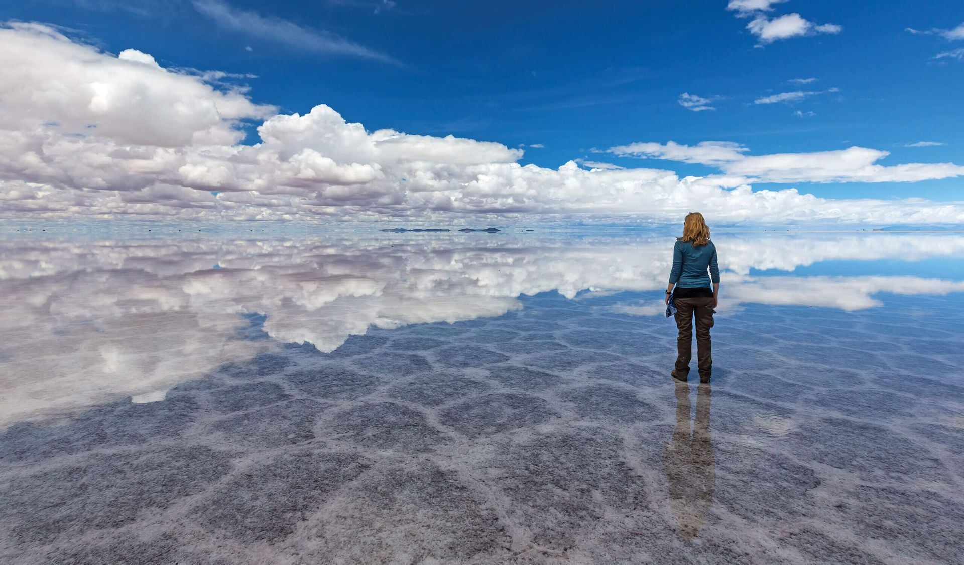 A person stands alone on a vast, reflective salt flat under a blue sky with scattered clouds—an unforgettable Bolivia travel scene, where the unique weather creates a stunning mirror-like effect on the ground.