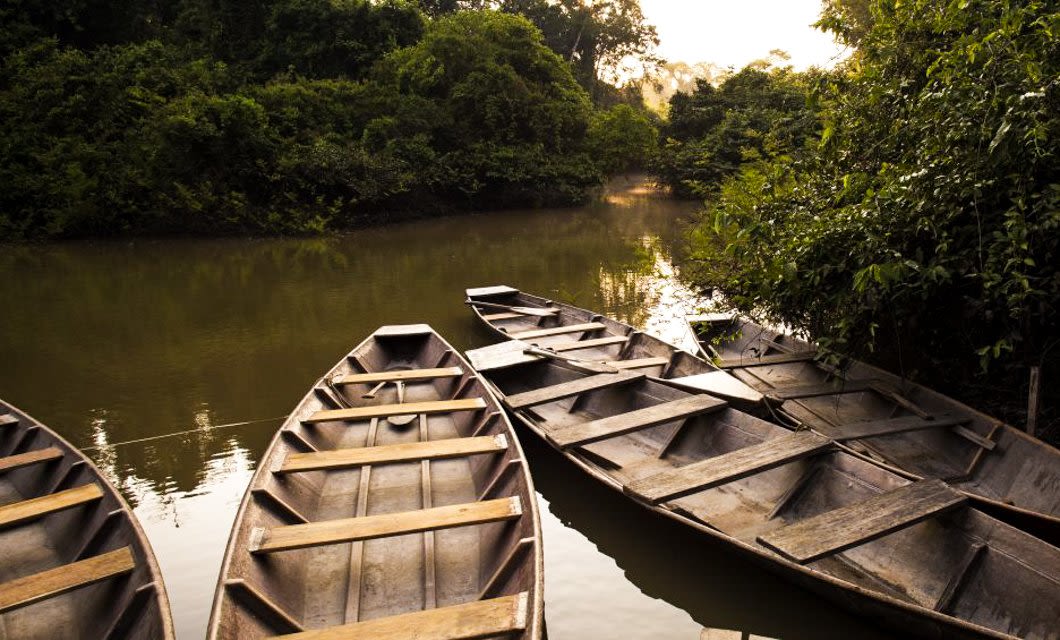 Five wooden boats are lined up along the edge of a calm, narrow river by Inkaterra Hacienda Concepción Lodge, surrounded by dense green trees and vegetation, with soft sunlight filtering through the foliage.