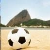 A soccer ball sits on a sandy beach with a large rocky hill and city buildings in the background under a clear sky, capturing the spirit of the World Cup 2024.