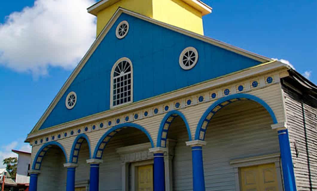 A colorful building with a blue and yellow facade, resembling the vibrant style of a Chiloe Hotel, features round windows and three blue columns supporting arched decorations above the entrance. The sky is partly cloudy in the background.