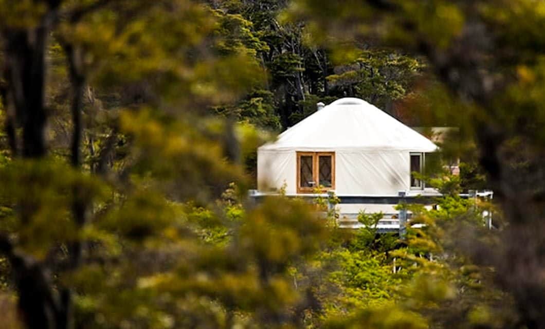 A white yurt with wooden-framed windows is nestled among green trees and foliage at Patagonia Camp, an eco-lodge in Patagonia, partially obscured by branches in the foreground.