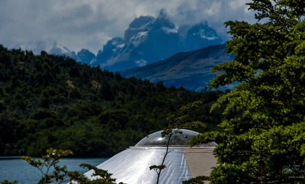 A silver dome-shaped structure, part of Patagonia Camp’s eco-friendly accommodation, is partially visible among green trees, with a lake and forested hills in the foreground and Torres del Paine’s snow-capped mountains under a cloudy sky in the background.