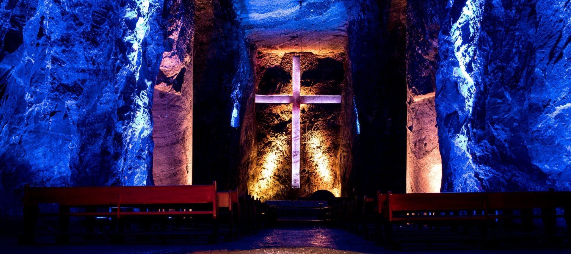 Underground chapel in Colombia with a large illuminated cross carved into a rock wall, glowing with warm light. The surrounding stone walls are lit with blue lights, and wooden pews are arranged in rows—perfect for UNESCO-inspired tours.