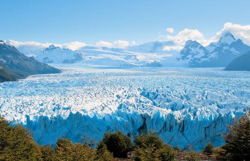 A vast glacier with jagged ice peaks stretches across the landscape, offering unforgettable experiences and activities amid snow-capped mountains, green trees, and a bright blue sky dotted with a few scattered clouds.
